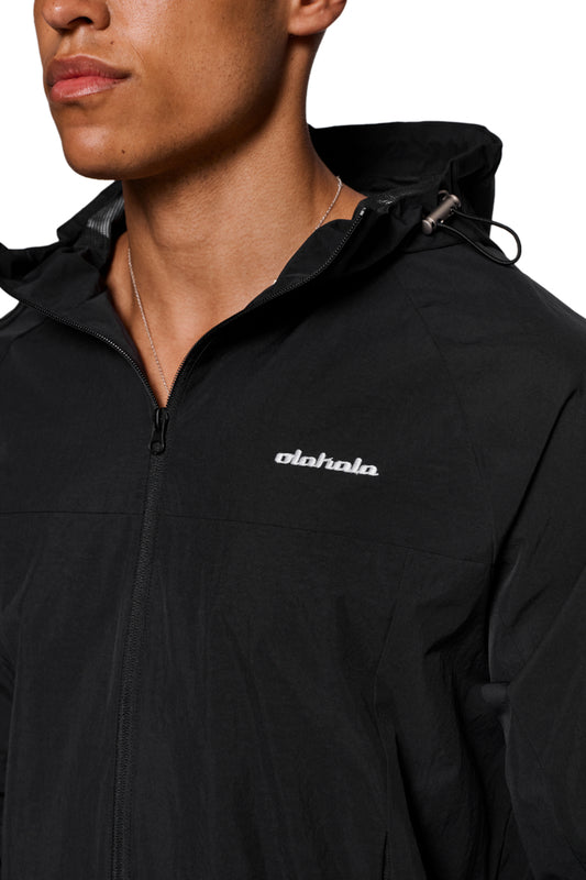 Person trägt Windbreaker Black mit verstellbarer Kapuze und OLAKALA-Stickerei links. Jacke aus wind- und wasserdichtem Material, elastische Ärmelbündchen und Reißverschlusstaschen sichtbar.